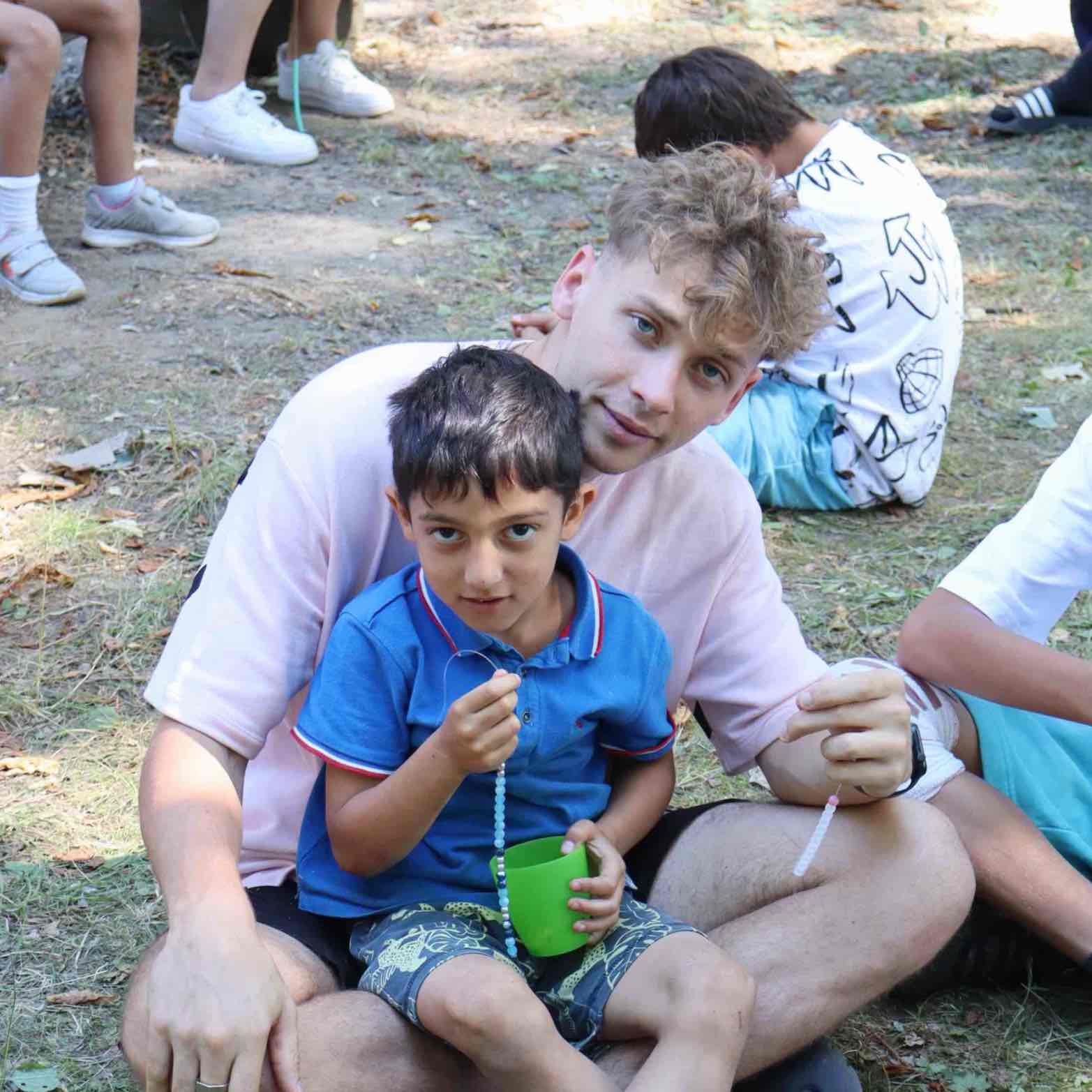 Vedoucí sedí s klukem na trávě a společně vyrábějí náramek
